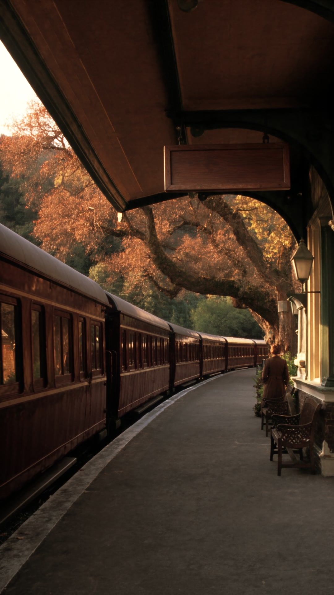 Chestnut-red train curving past a heritage station beneath autumn trees